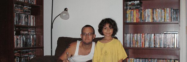 Uncle sitting in oversized chair with young nephew standing next to chair and overflowing bookcases containing books and movies.