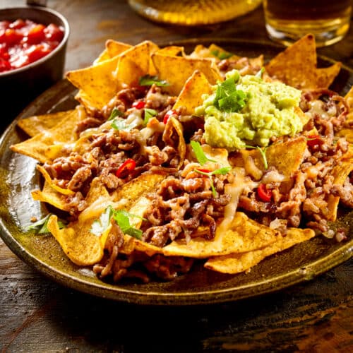 Yellow corn nacho chips garnished with ground beef, guacamole, melted cheese, peppers and cilantro leaves in plate on wooden table